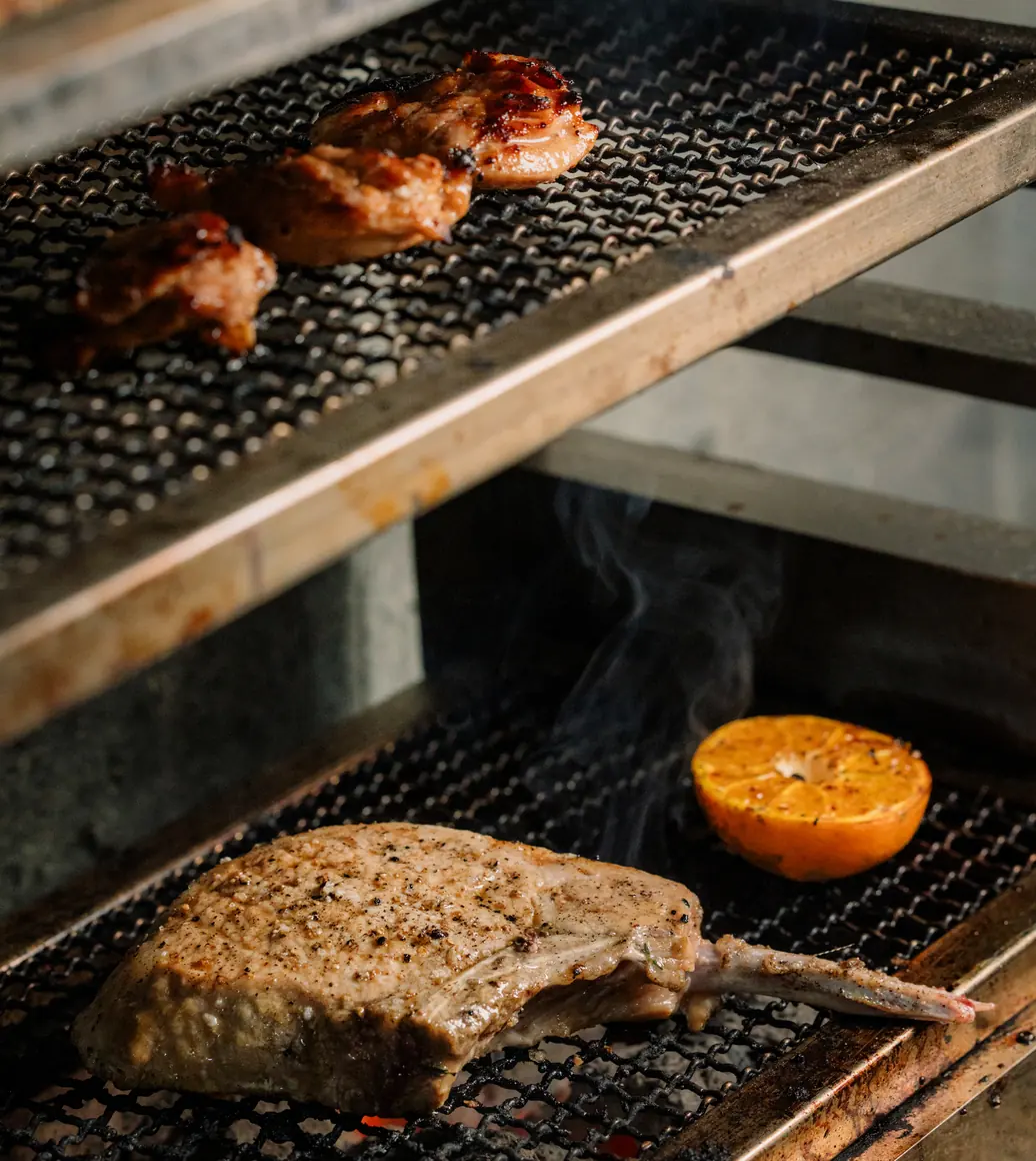 Meat and a grilled citrus half cooking over binchotan charcoal on the robata grill